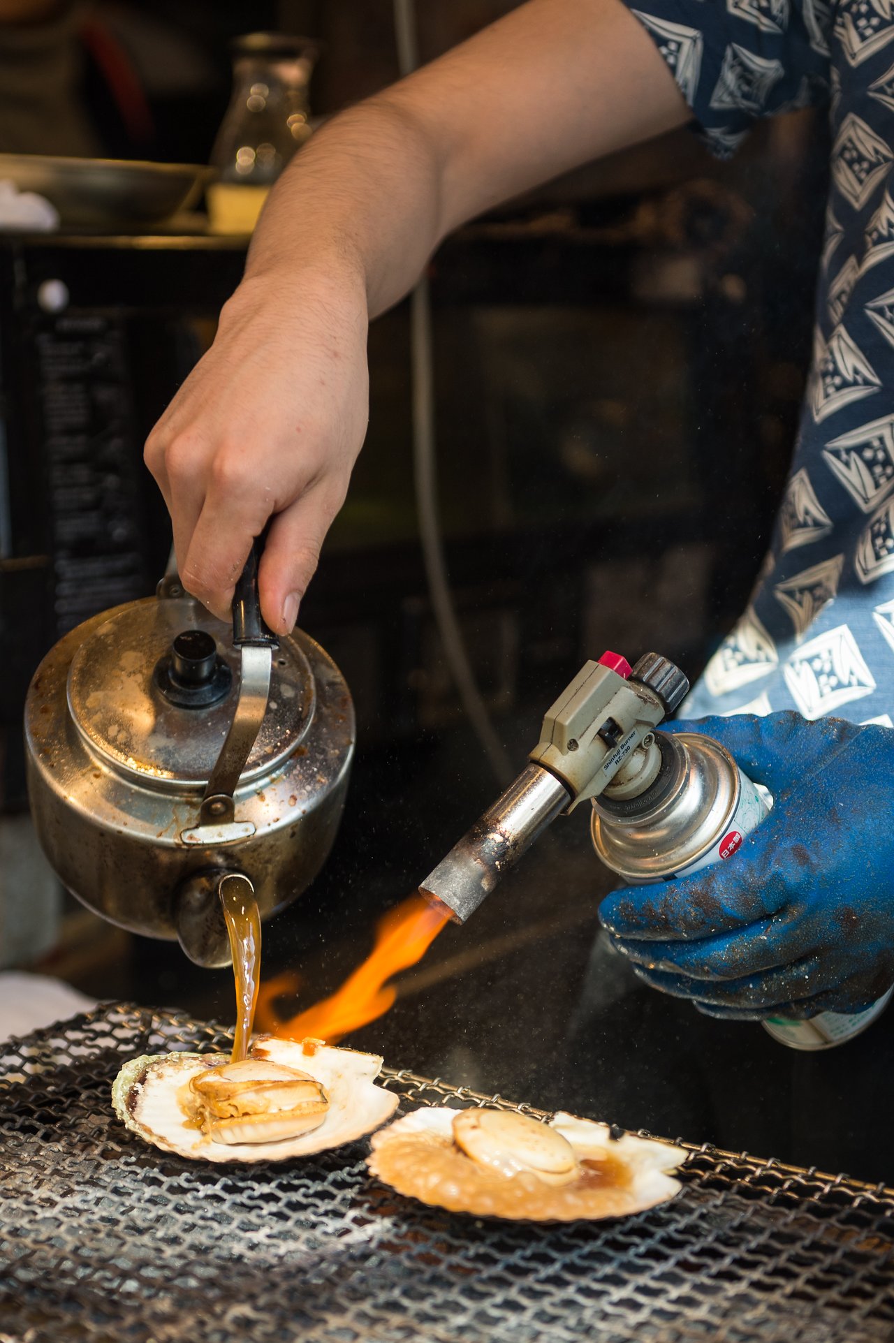 A person grills scallops with a blowtorch while pouring sauce from a kettle onto the seafood.