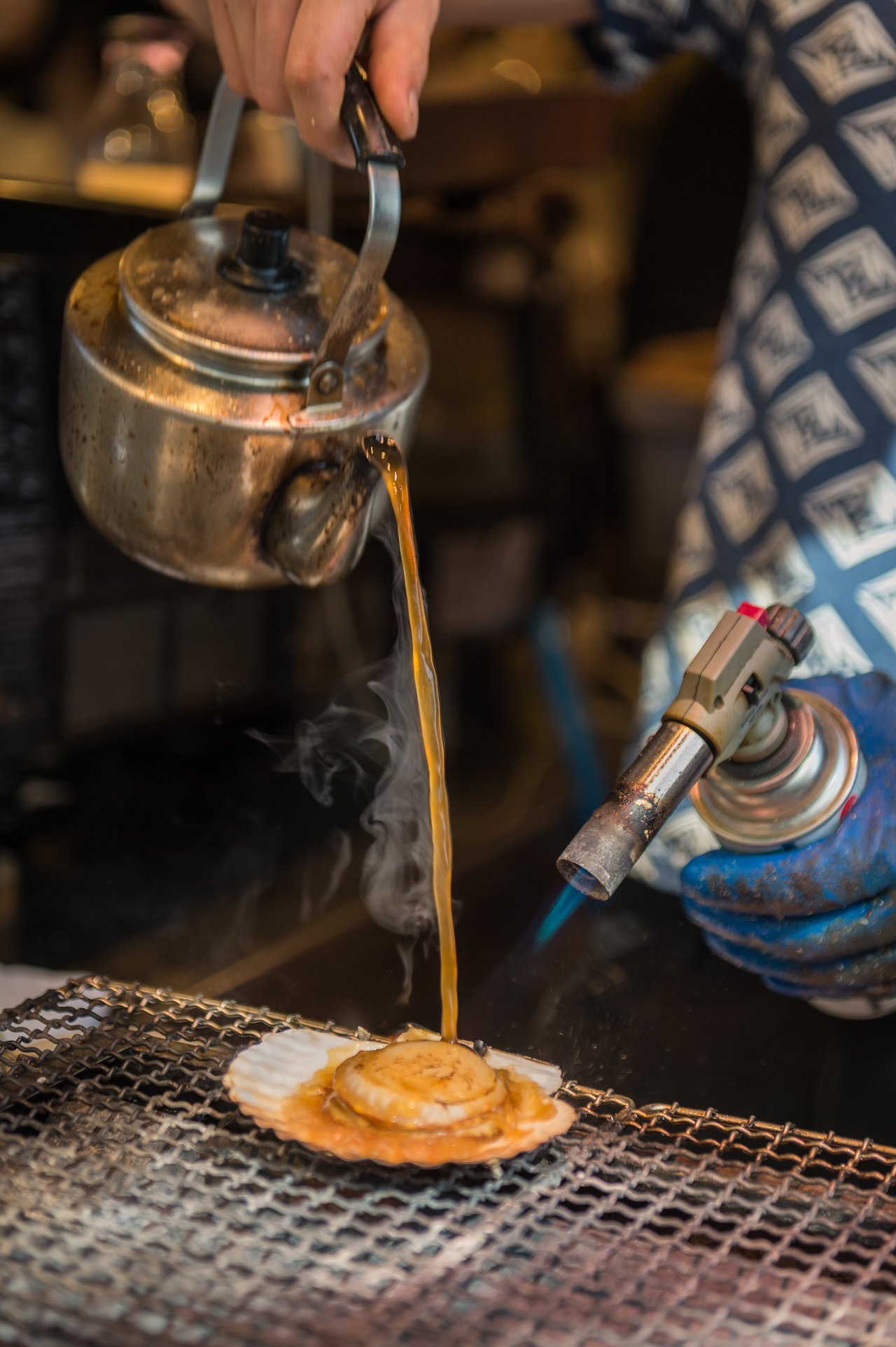 A person pours liquid from a kettle onto a scallop while using a blowtorch to cook it.