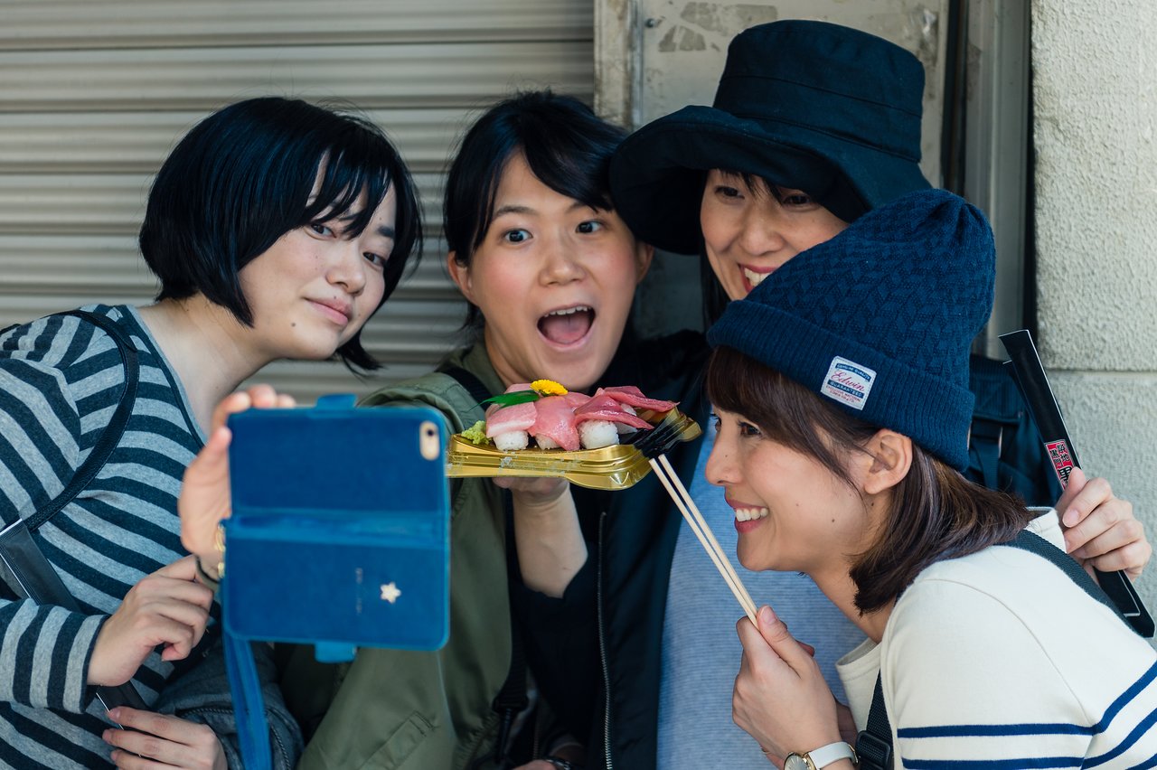 Four friends take a selfie while holding a tray of sushi, smiling and posing together.