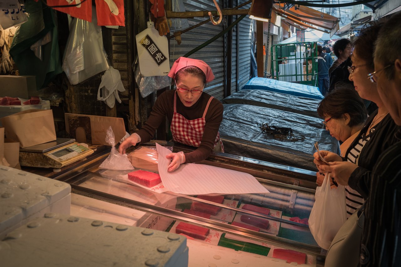 A vendor in a red apron wraps a piece of fish while customers wait at a market stall.