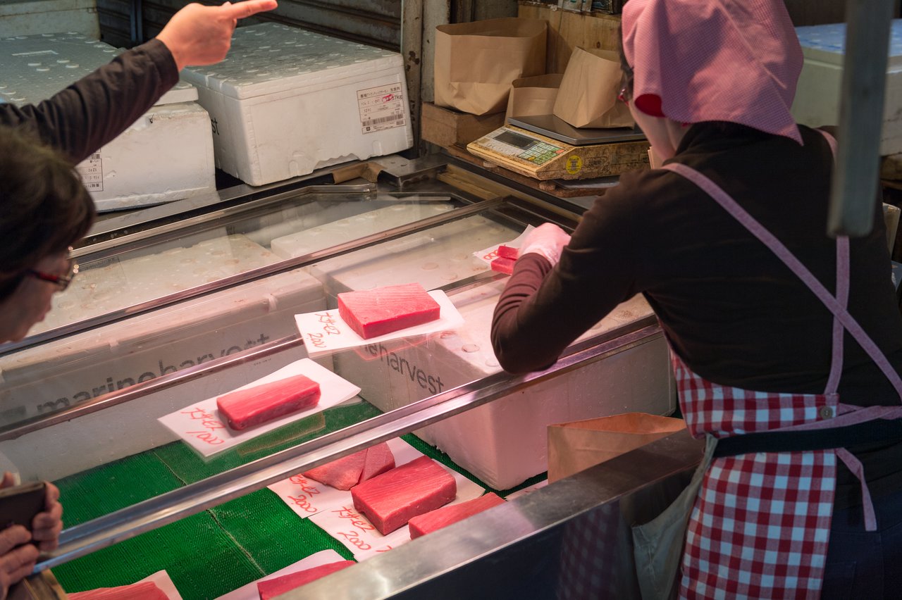A vendor in a red apron wraps a piece of tuna for a customer.
