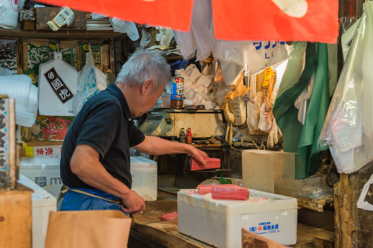 A fish vendor in a market cuts and handles fresh tuna on a wooden counter with styrofoam boxes nearby.