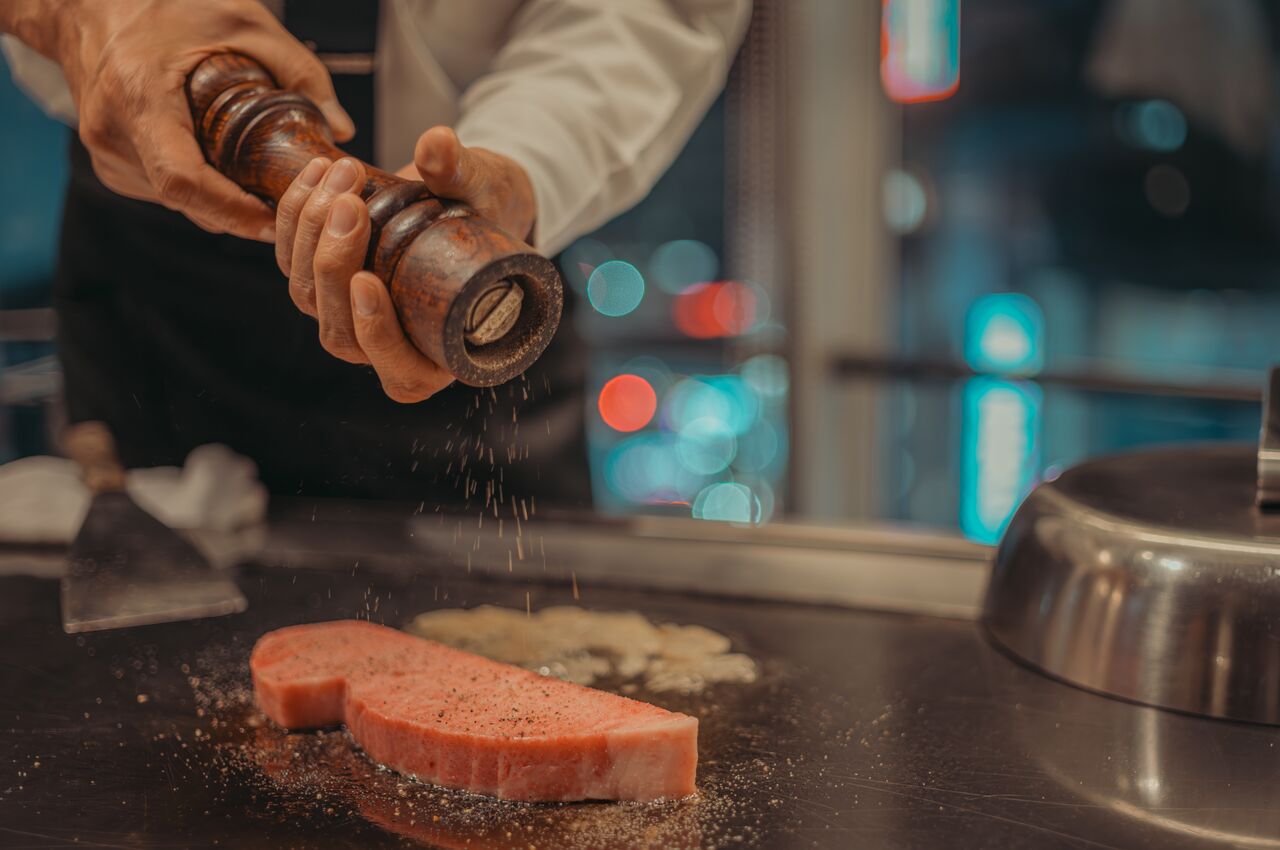 A chef seasons a thick slice of raw Kobe beef with pepper on a hot teppanyaki grill at a restaurant in Kobe, Japan.