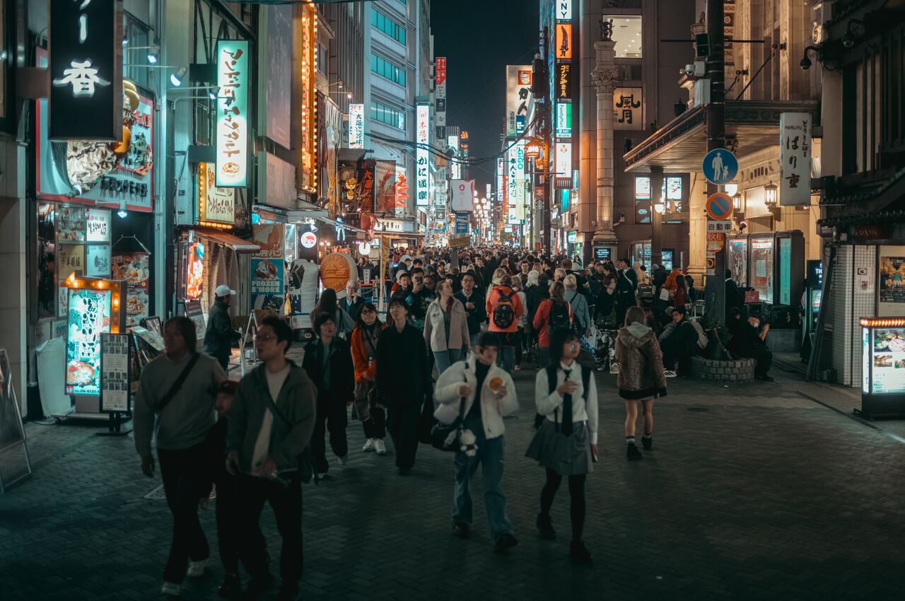 A large crowd walks through a brightly lit street in Dotonbori at night, surrounded by restaurants and colorful signs.