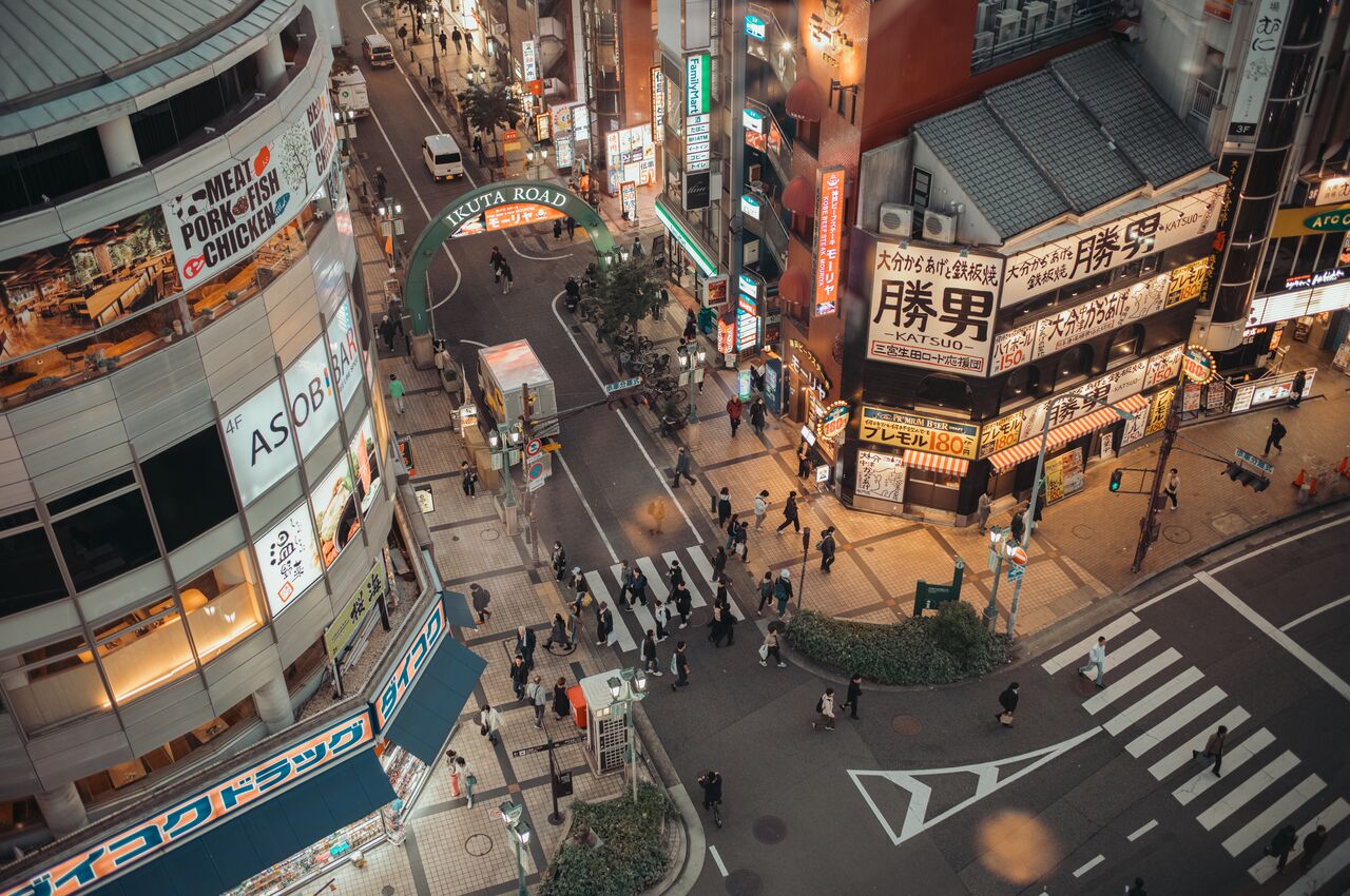 People walk through a busy intersection surrounded by brightly lit buildings and shops in an urban area of Kobe, Japan.