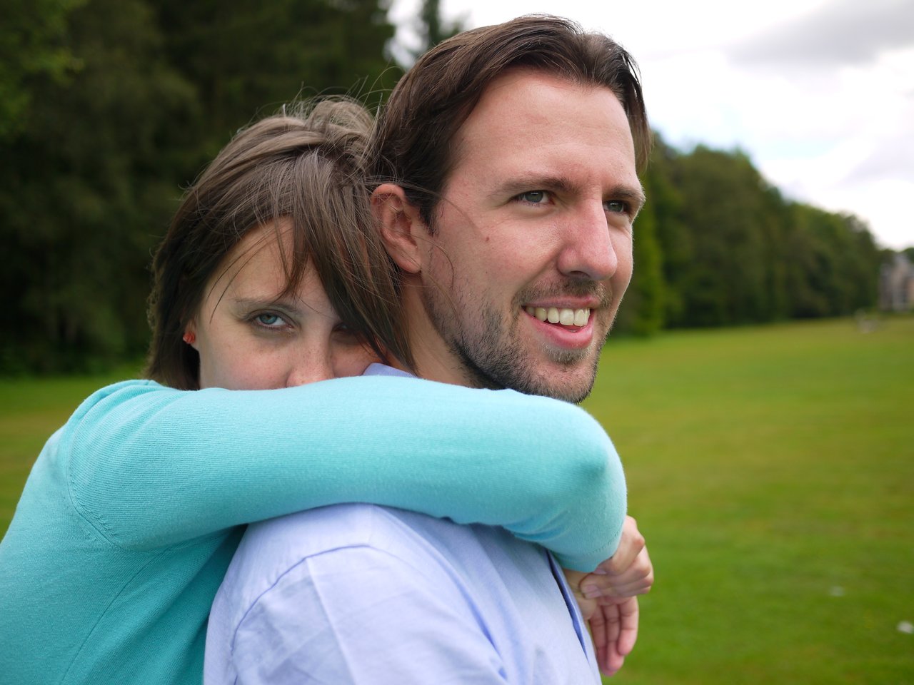 A woman in a blue sweater hugs a smiling man from behind in an open grassy area.