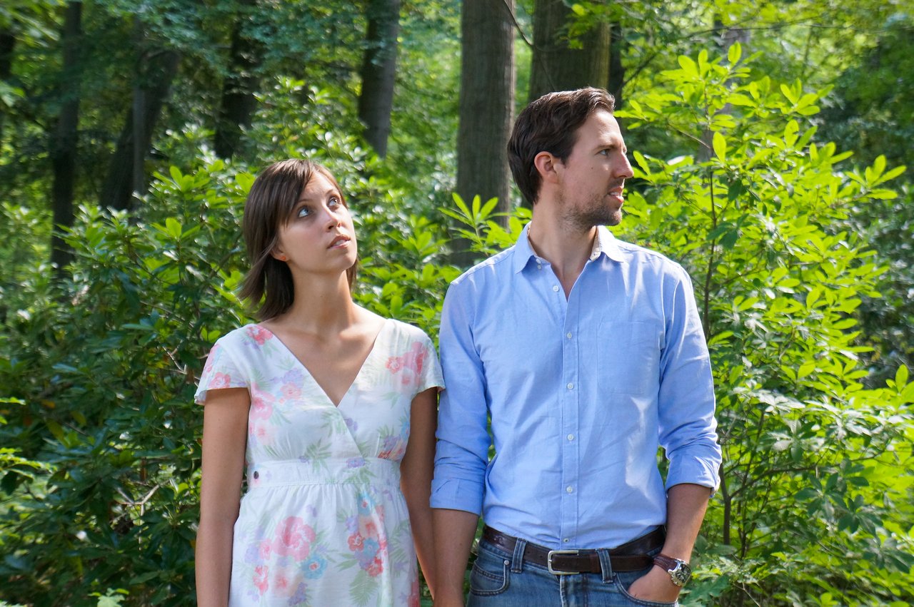 A man and woman stand outdoors holding hands, looking in different directions with neutral expressions.