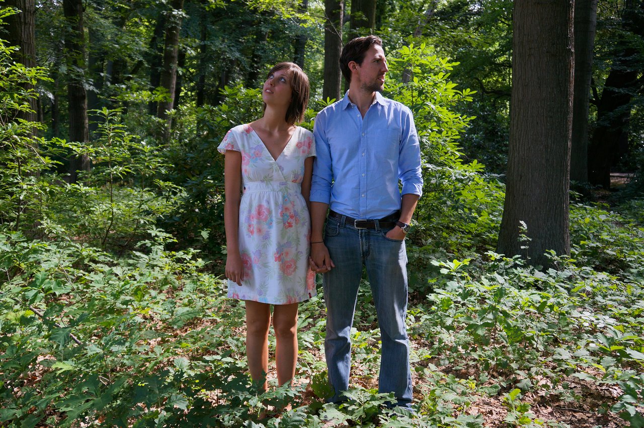 A man and woman stand in a forest holding hands, looking in different directions with neutral expressions.