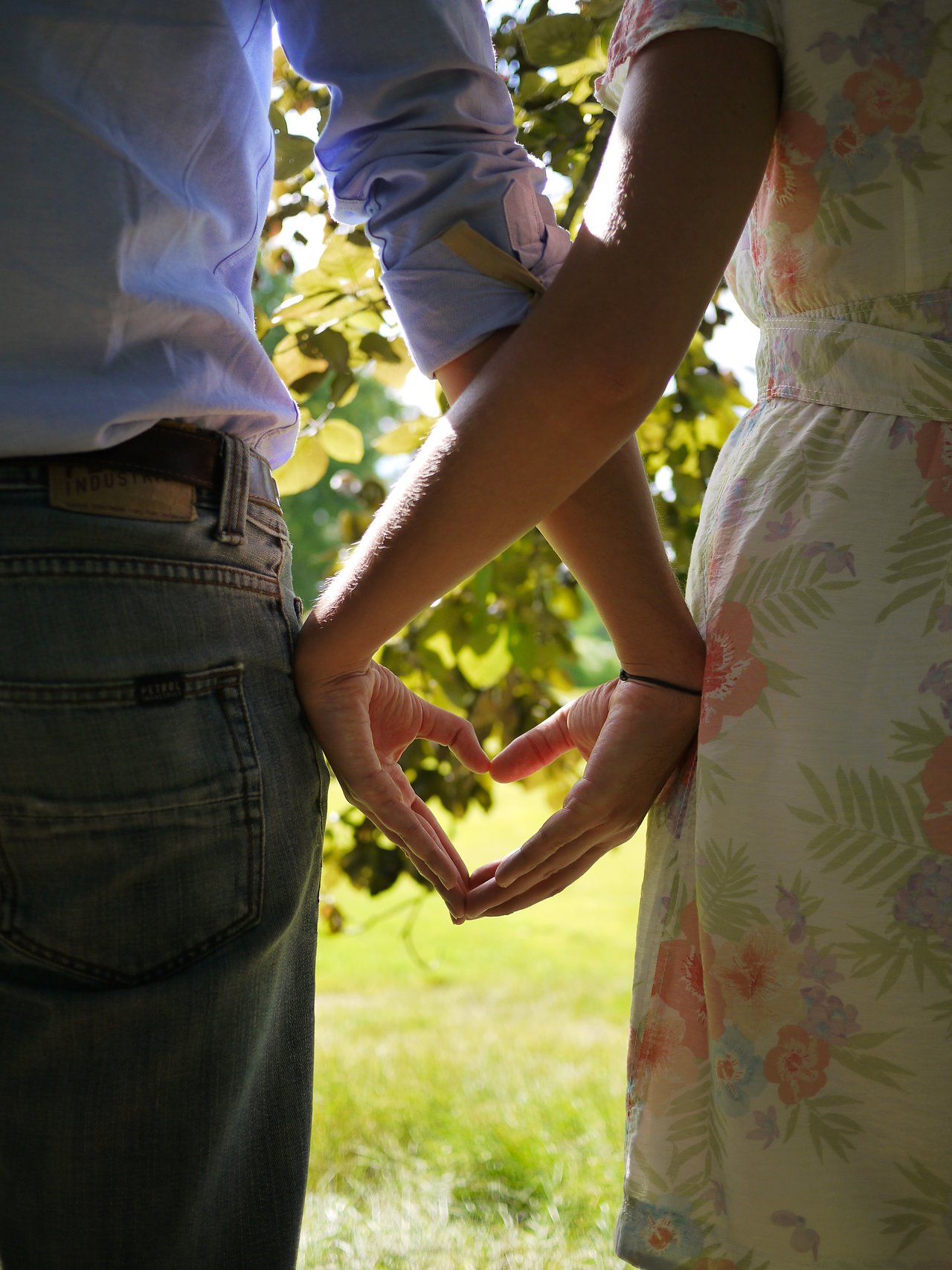 A couple stands side by side, forming a heart shape with their hands against a sunlit outdoor background.