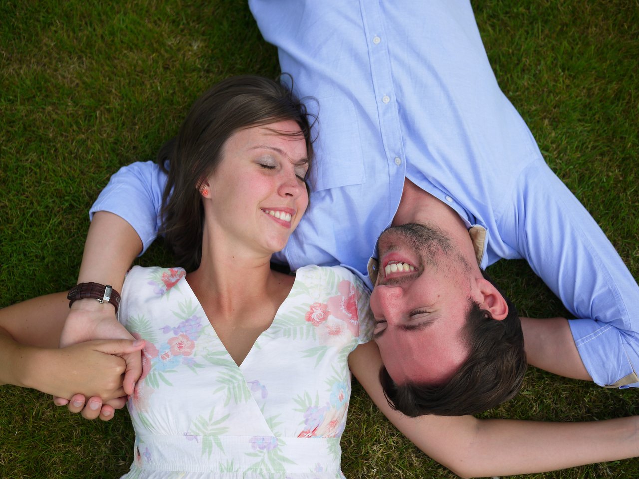 A smiling couple lies on the grass, holding hands and looking happy together.