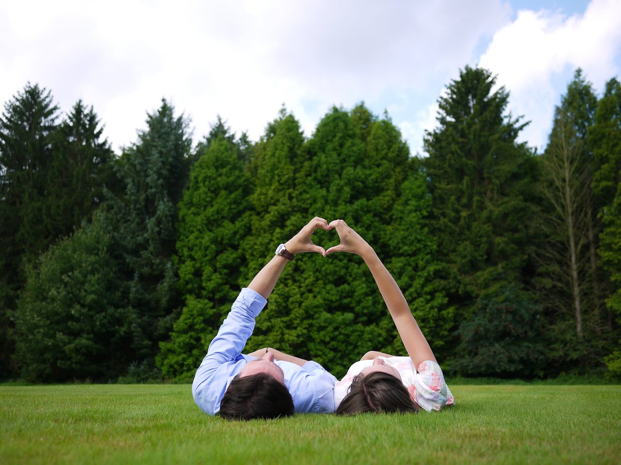 Two people lying on the grass form a heart shape with their hands against a backdrop of green trees.