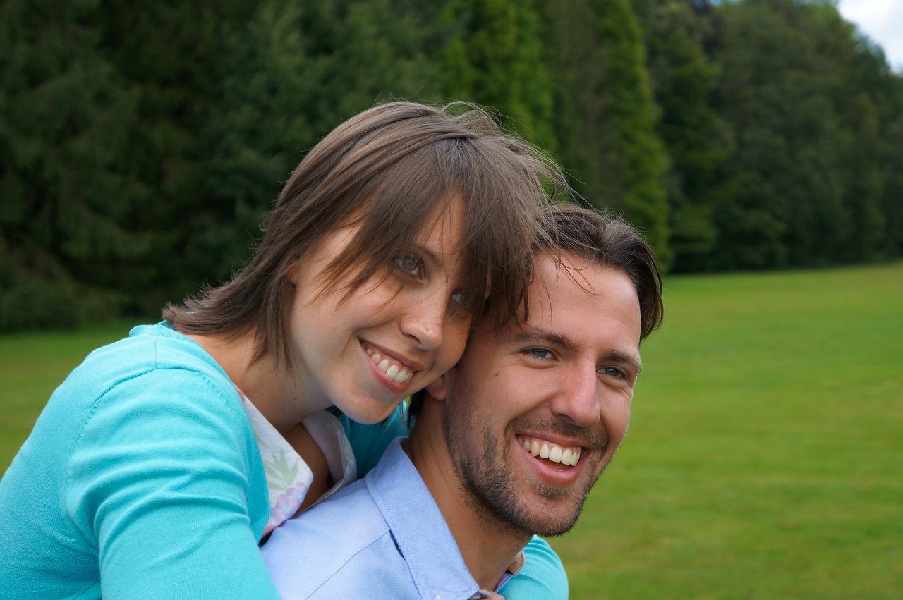 A smiling woman leans on a smiling man's back in an outdoor setting with green grass and trees.