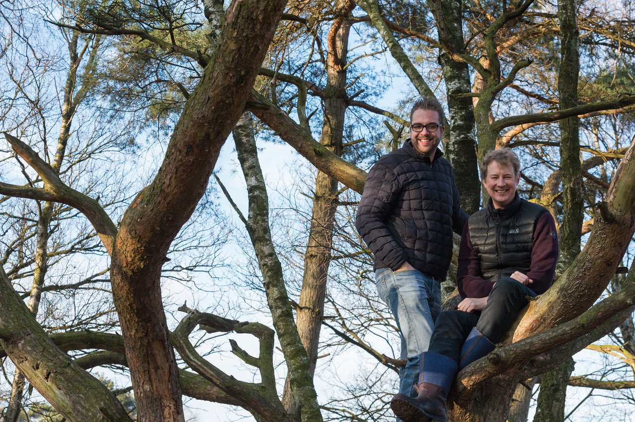 Two people wearing jackets sit on the branches of a tree, smiling at the camera in a wooded area.