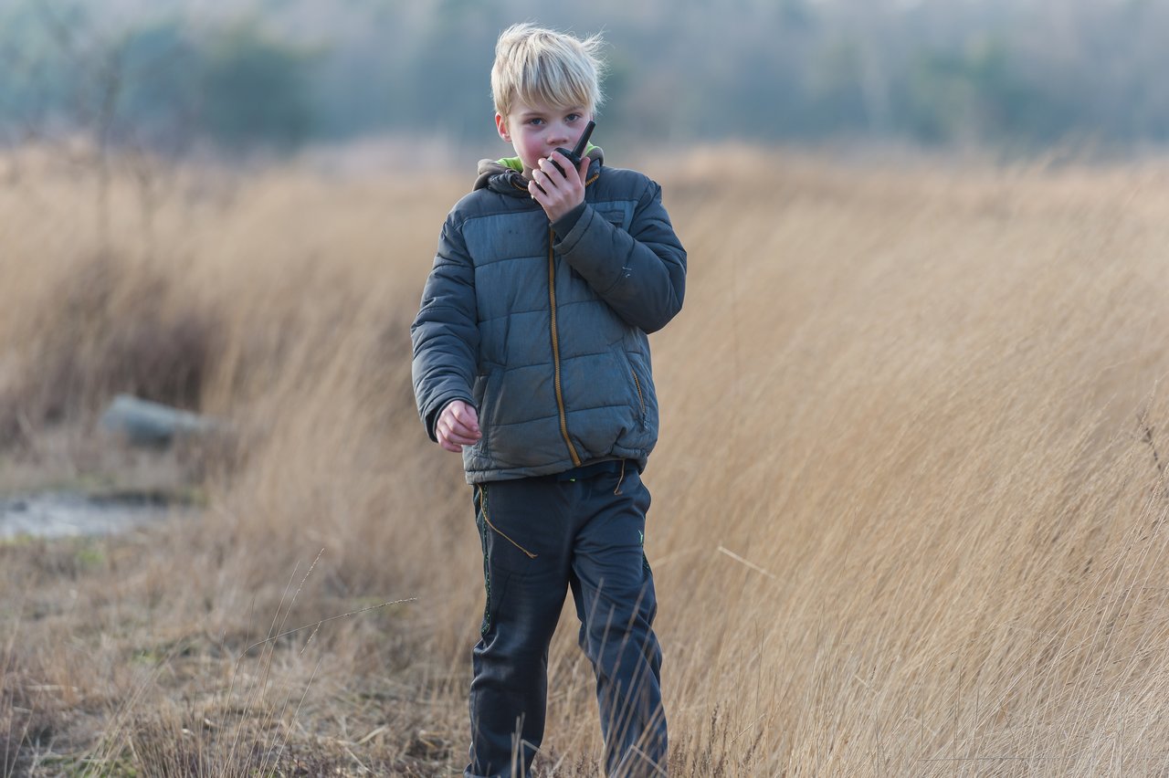A child in a winter jacket walks through tall grass while speaking into a walkie-talkie.