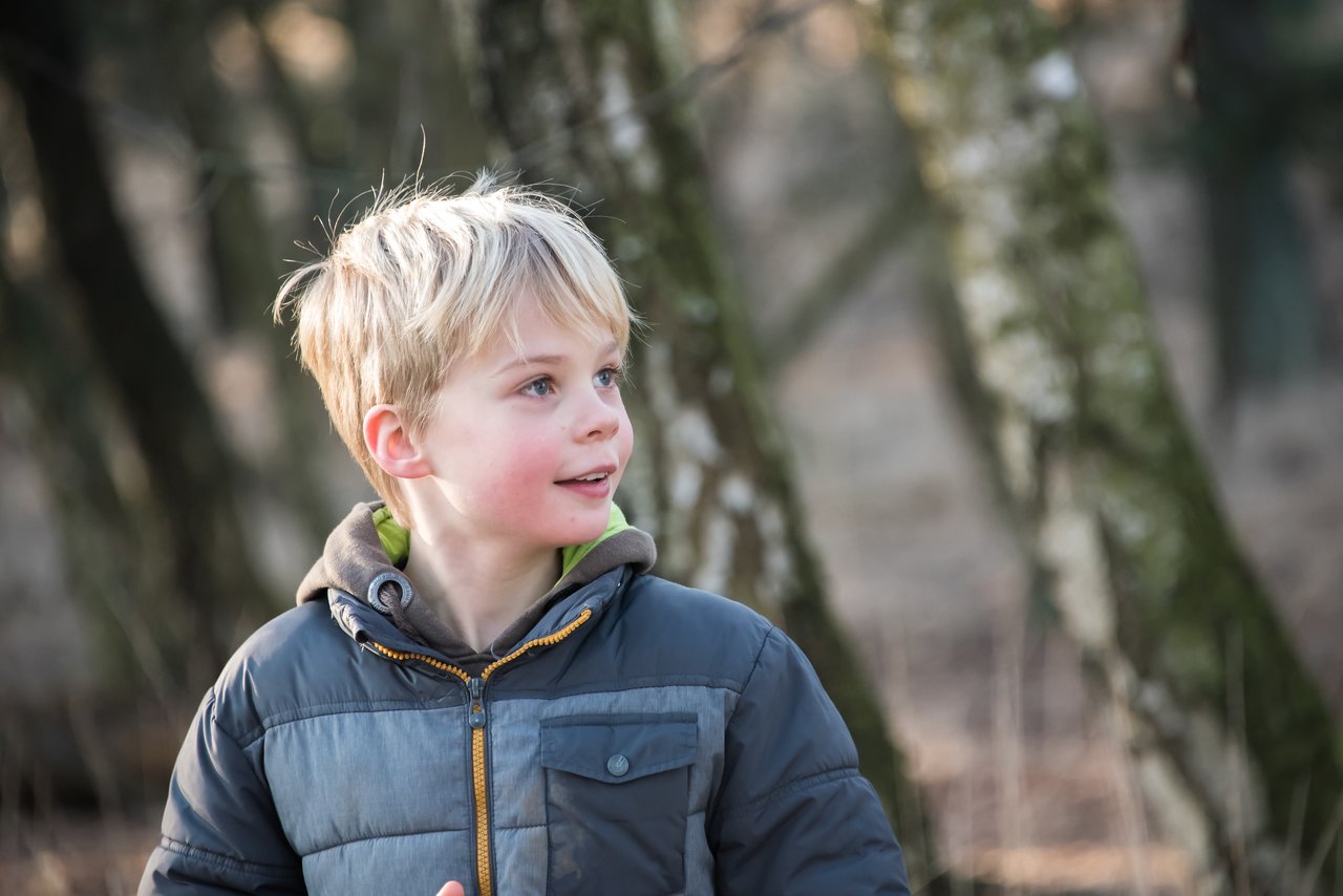 A young boy in a winter jacket looks to the side while standing in a wooded area.
