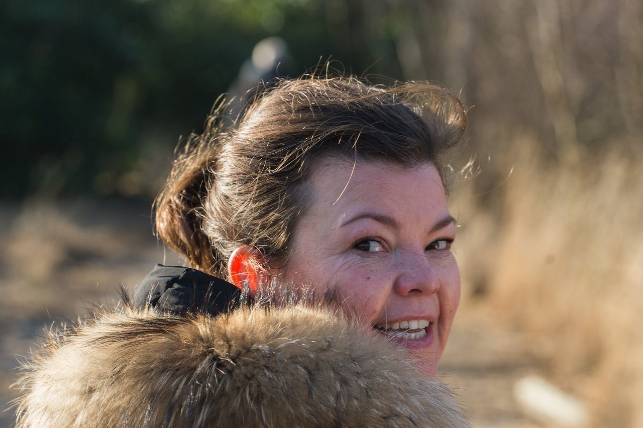 A woman with a fur-lined coat smiles while looking back on a sunlit outdoor trail.