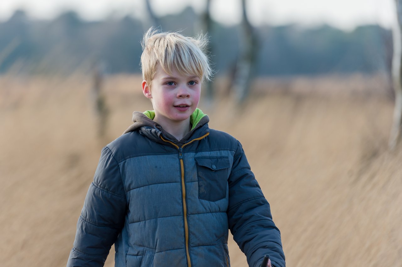 A young child in a winter jacket walks outdoors in a grassy field with trees in the background.