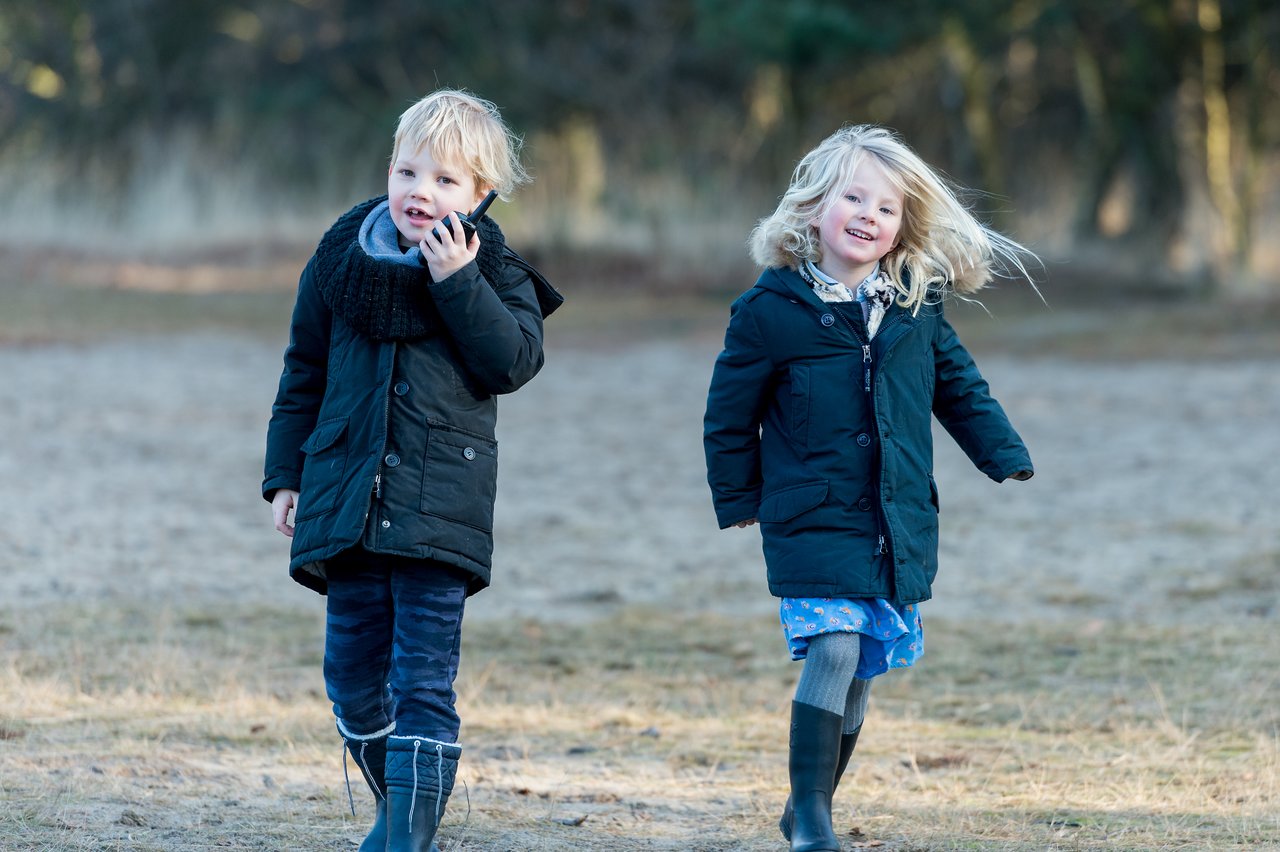 Two children in warm coats and boots walk outdoors on a sandy path, one smiling and the other touching their face.