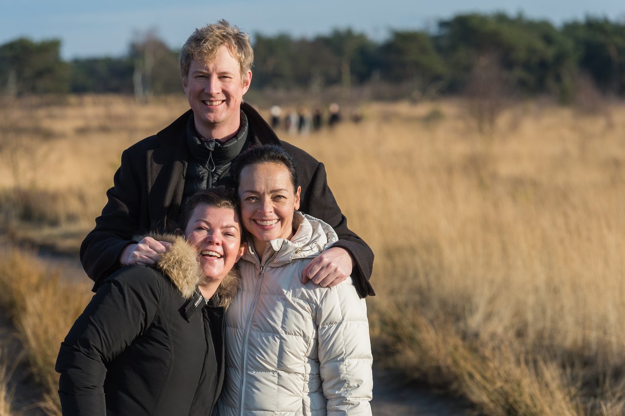 Three people in warm jackets smile and pose together outdoors in a grassy landscape on a sunny day.