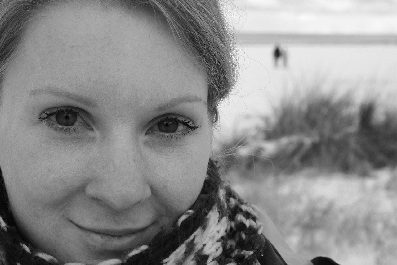Close-up of a woman with a scarf, smiling slightly, with a blurred beach and distant figures in the background.
