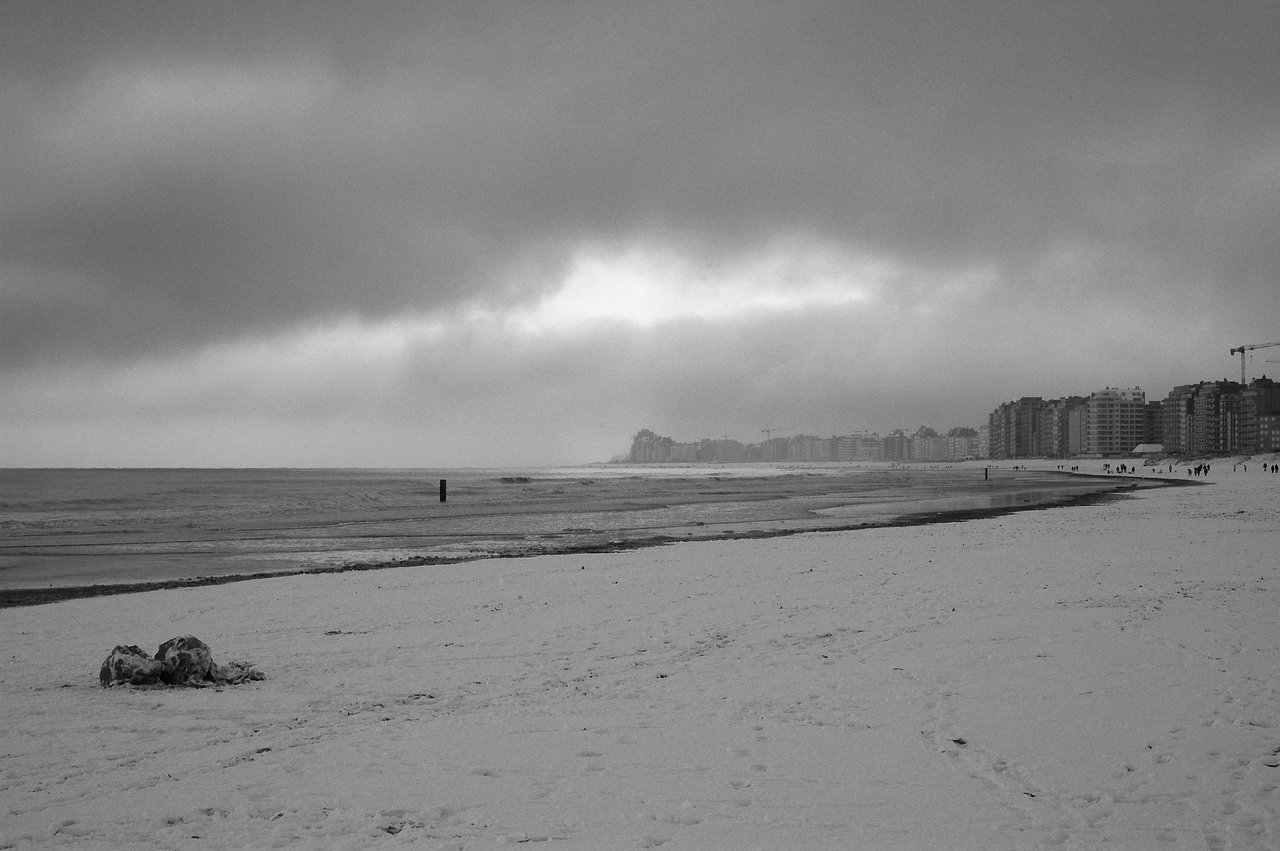 A snowy beach with footprints, calm waves, and a distant city skyline under a cloudy sky.