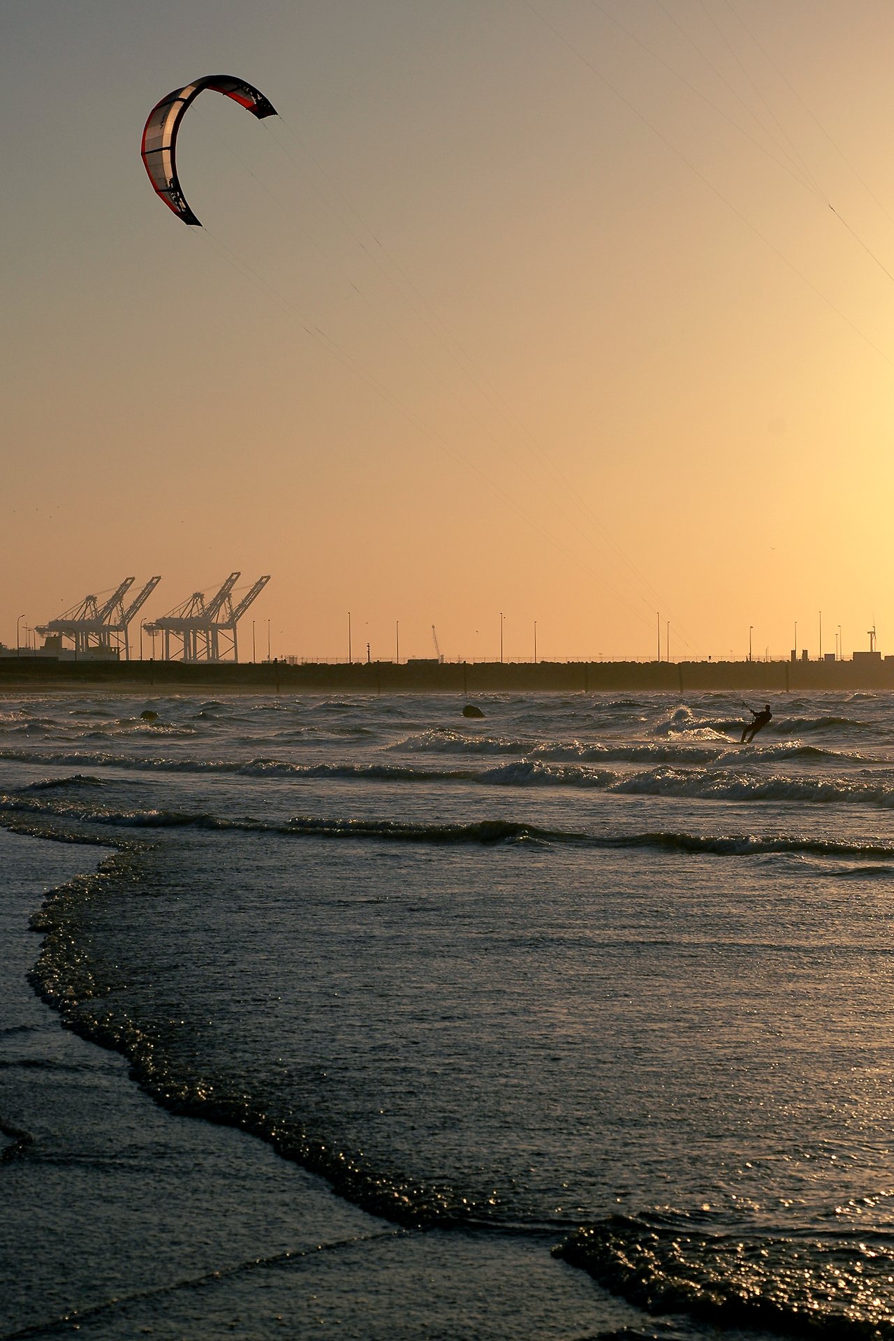 A kite surfer glides over the waves near the shore, with a large kite flying high above.