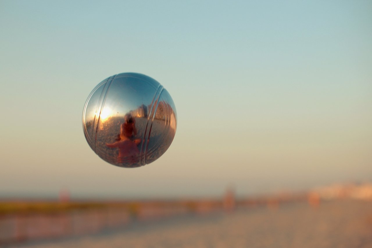 A pétanque ball is captured mid-air, reflecting the photographer and the beach in its shiny surface.
