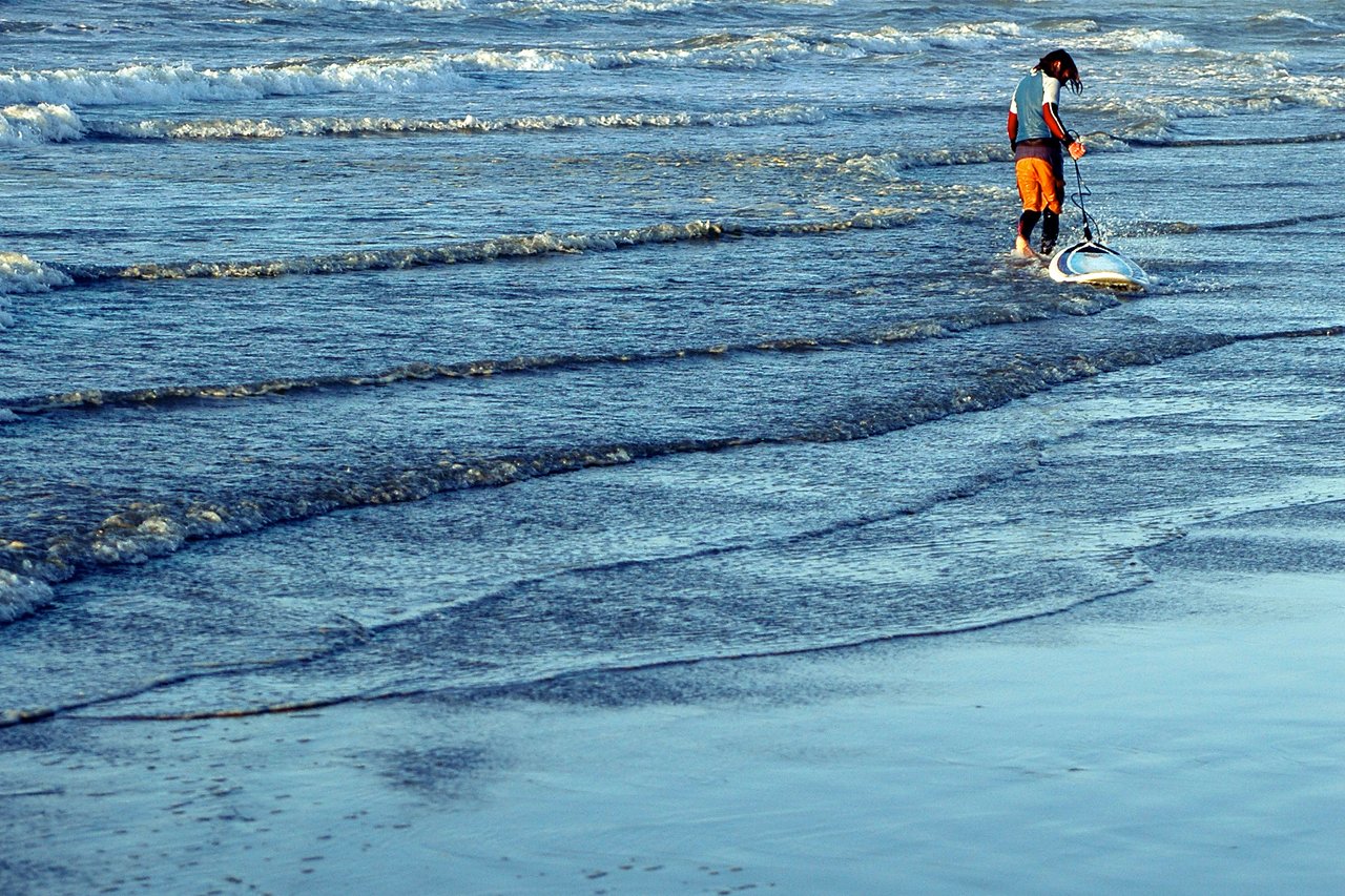 A person in a wetsuit and shorts pulls a surfboard through shallow water near the shore.
