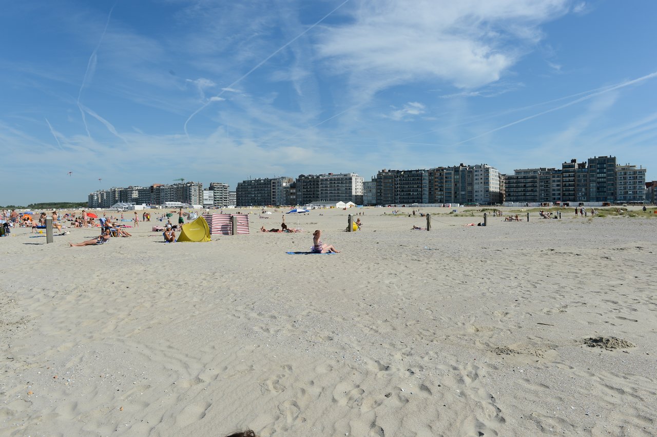 People are relaxing on a sandy beach with umbrellas and tents, with large apartment buildings in the background.