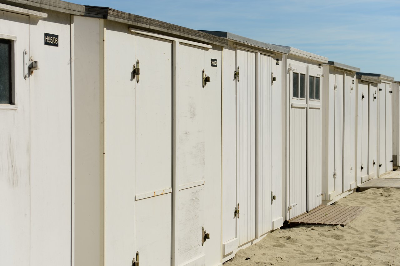 A row of white beach cabins with closed doors, standing on sandy ground under a clear blue sky.