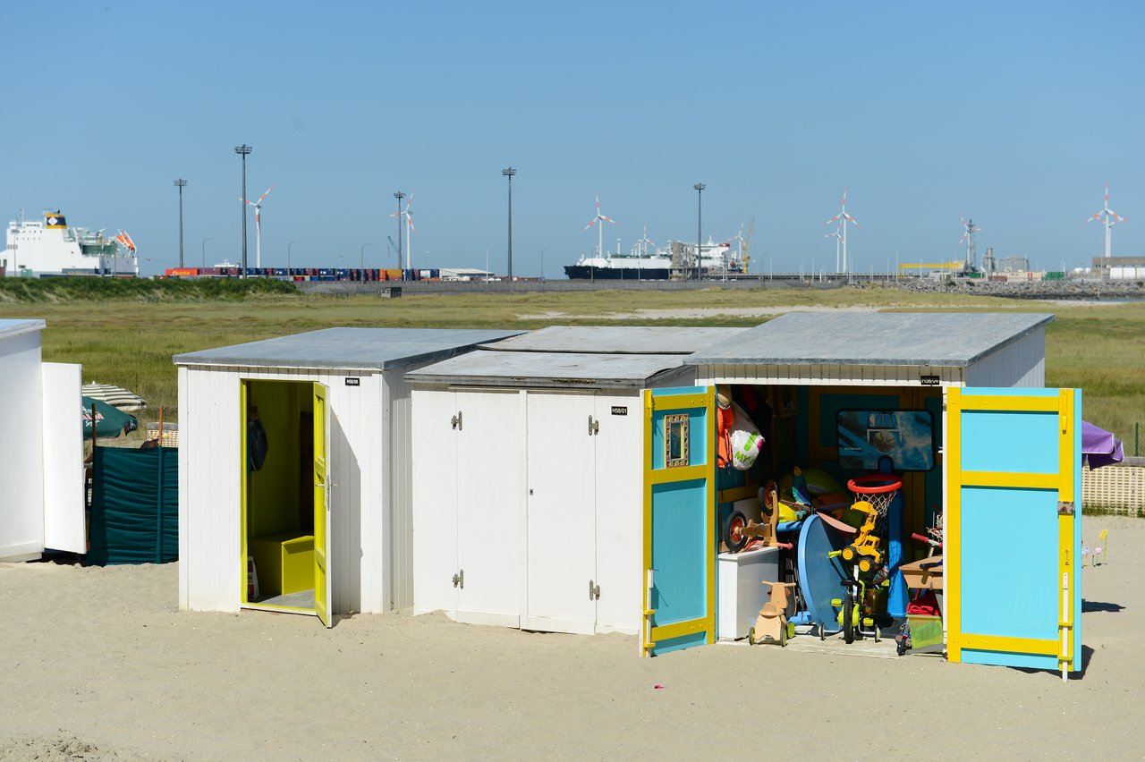 White beach cabins with open doors, one revealing storage with toys and beach gear, set on sandy ground.