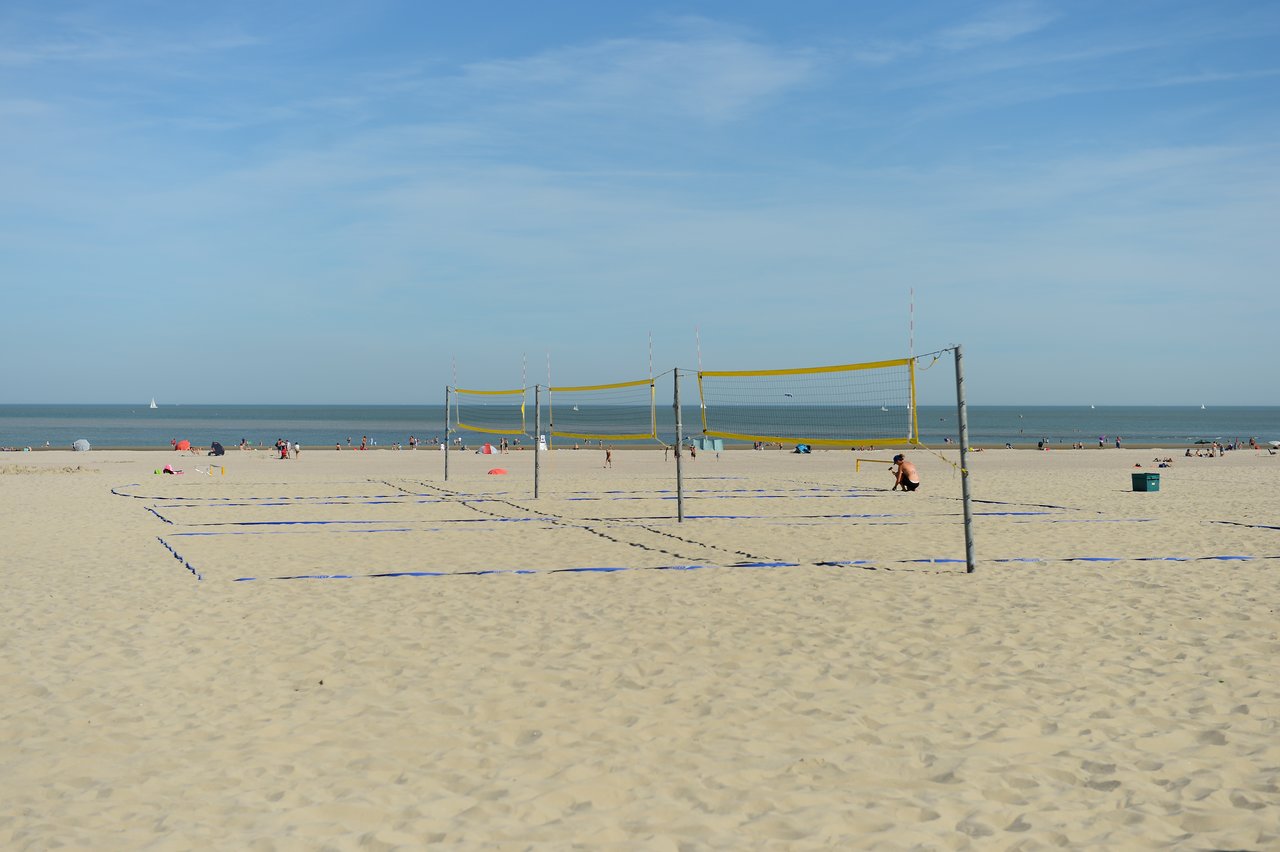 A beach volleyball court with yellow nets, and a person sitting on the sand near the net.