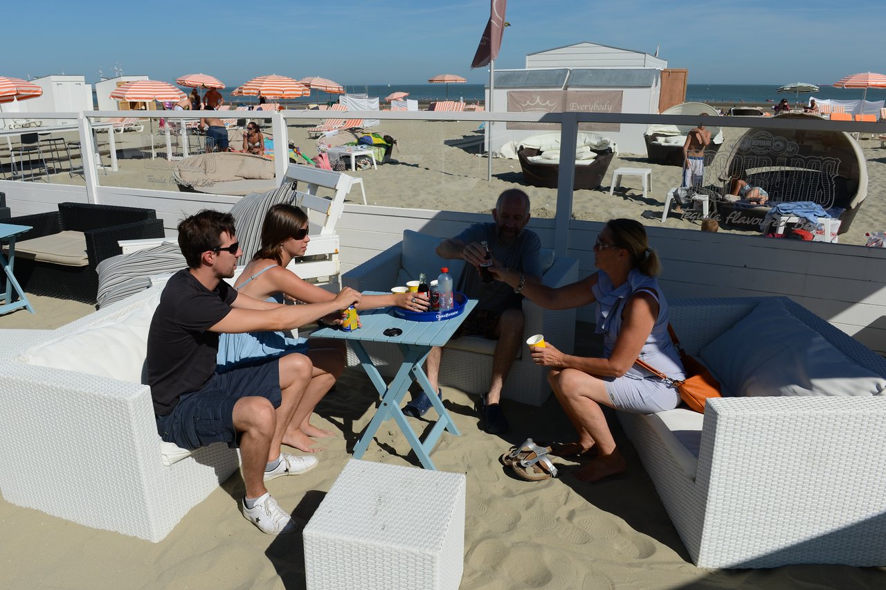 Four people sit on white outdoor couches at a beachside café, raising their drinks in a toast.