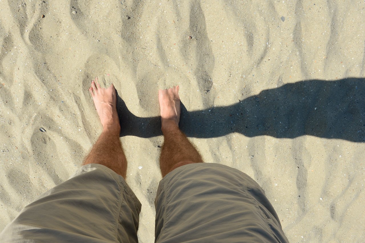A person stands barefoot on sandy ground, wearing khaki shorts, with a shadow stretching forward.