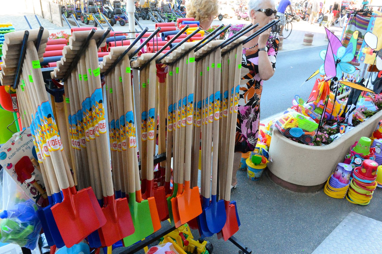 A display of colorful shovels with wooden handles at an outdoor market, with two women browsing nearby.