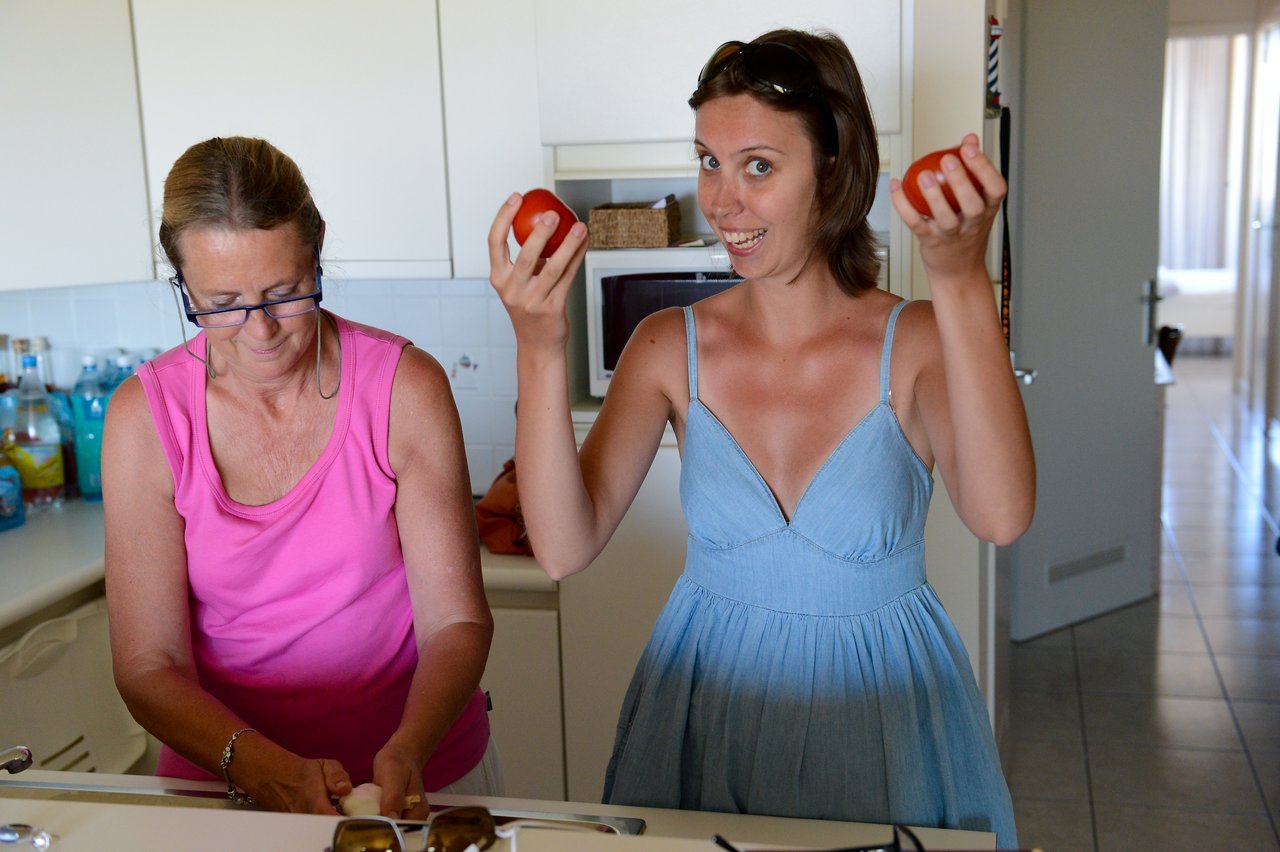Two women in a kitchen, one cutting food while the other smiles and holds up two tomatoes.
