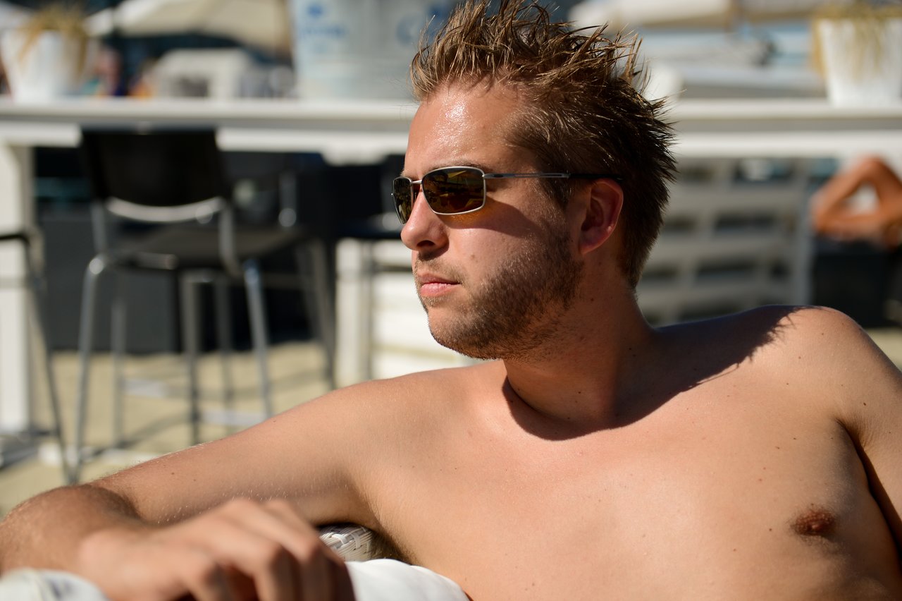 A man wearing sunglasses lounges shirtless in the sun, looking off to the side with a relaxed expression.