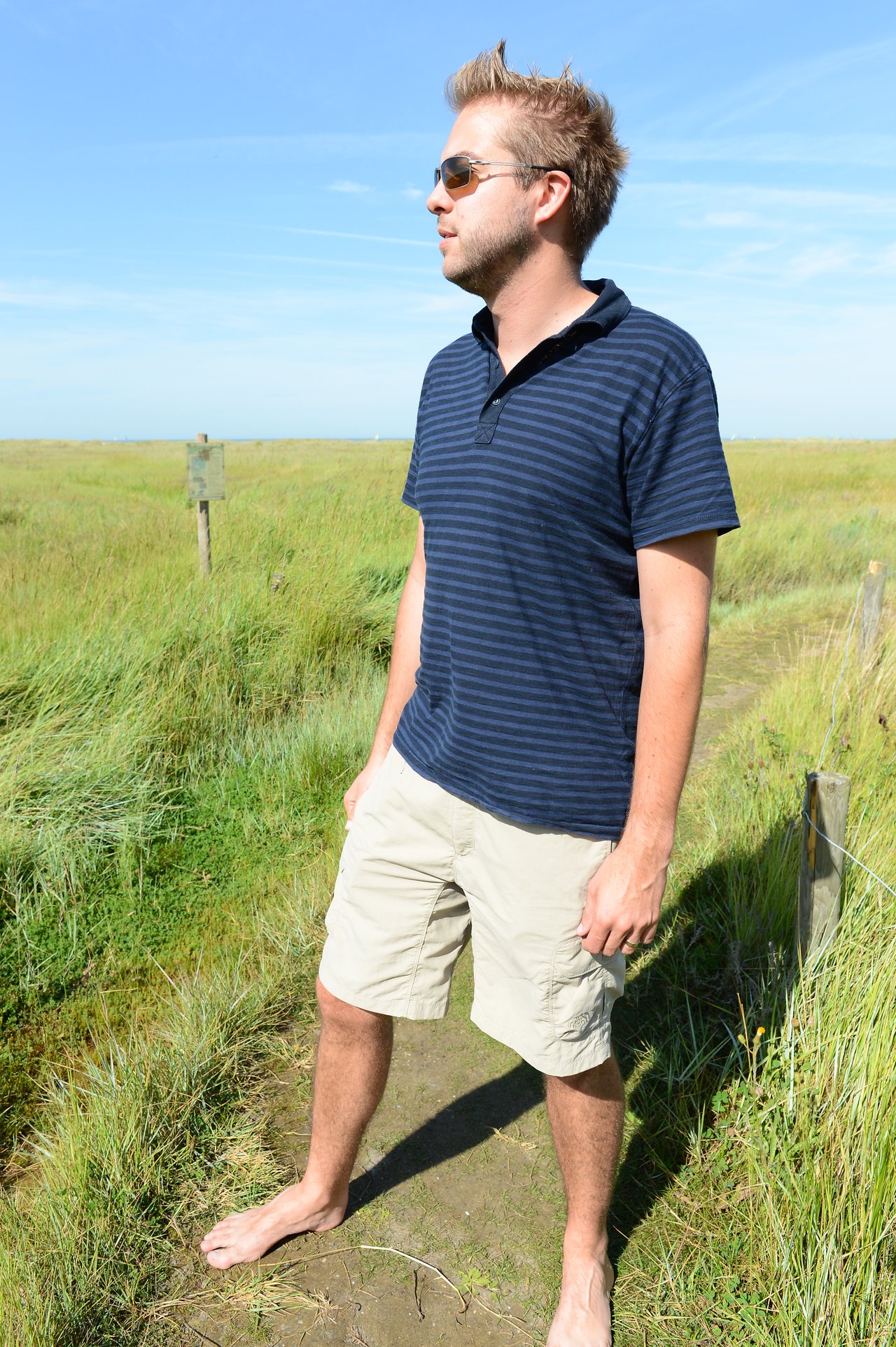 A man wearing sunglasses and casual clothes stands barefoot on a narrow dirt path in a grassy field.
