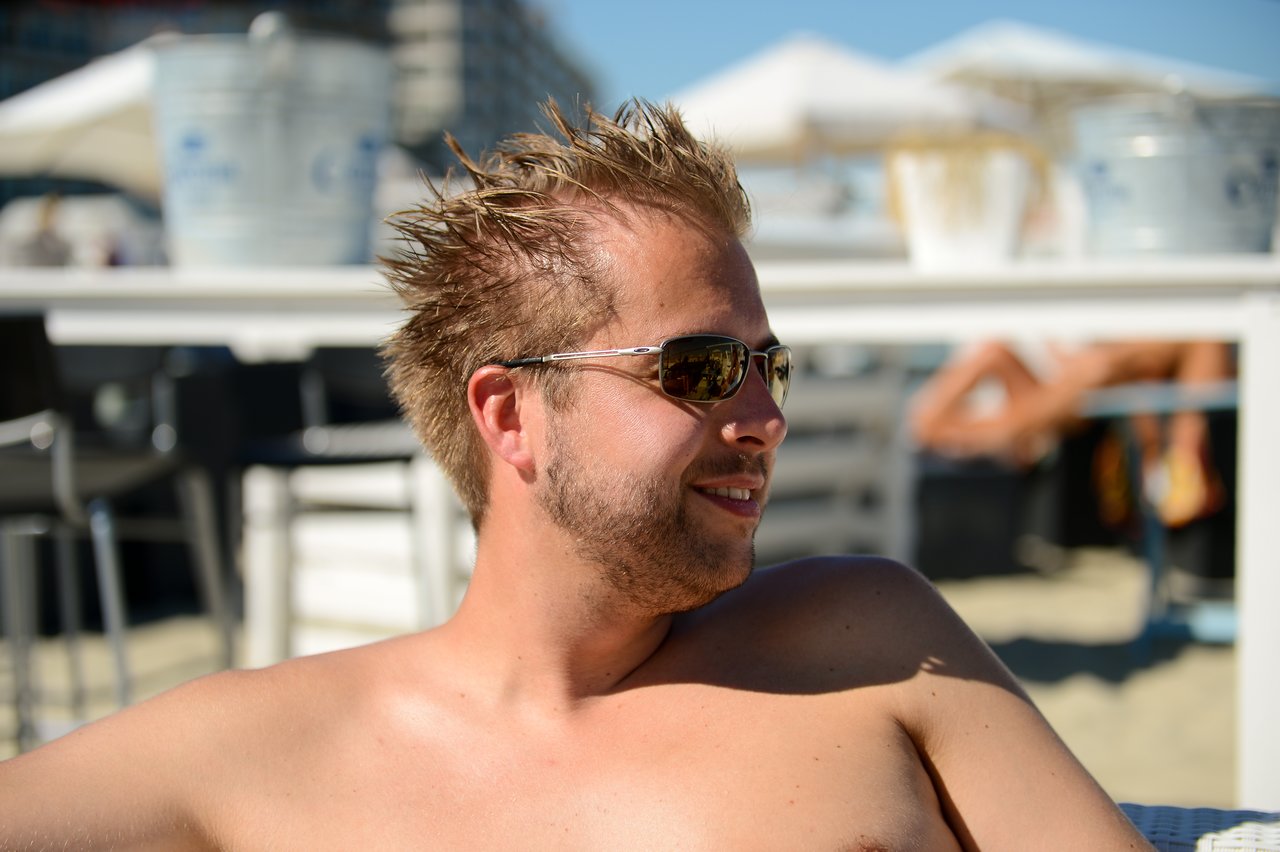 A man with spiky hair and sunglasses smiles while relaxing shirtless at a beachside location.