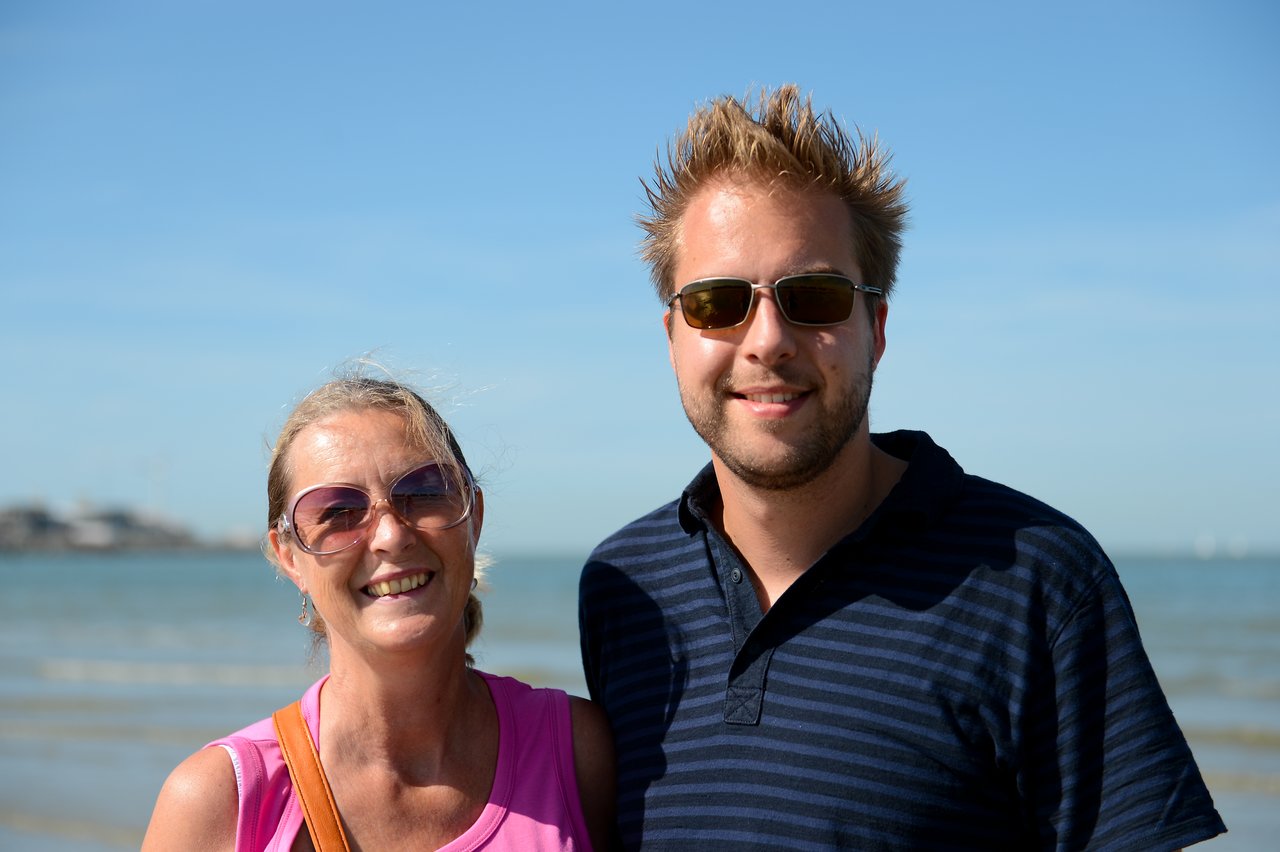 A man and a woman wearing sunglasses smile while posing together on a sunny beach.