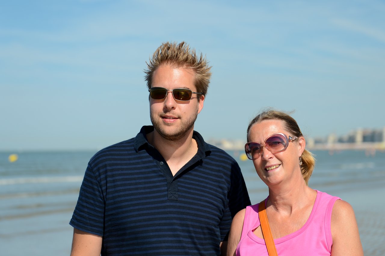 A man and a woman wearing sunglasses stand together on a beach, smiling at the camera.