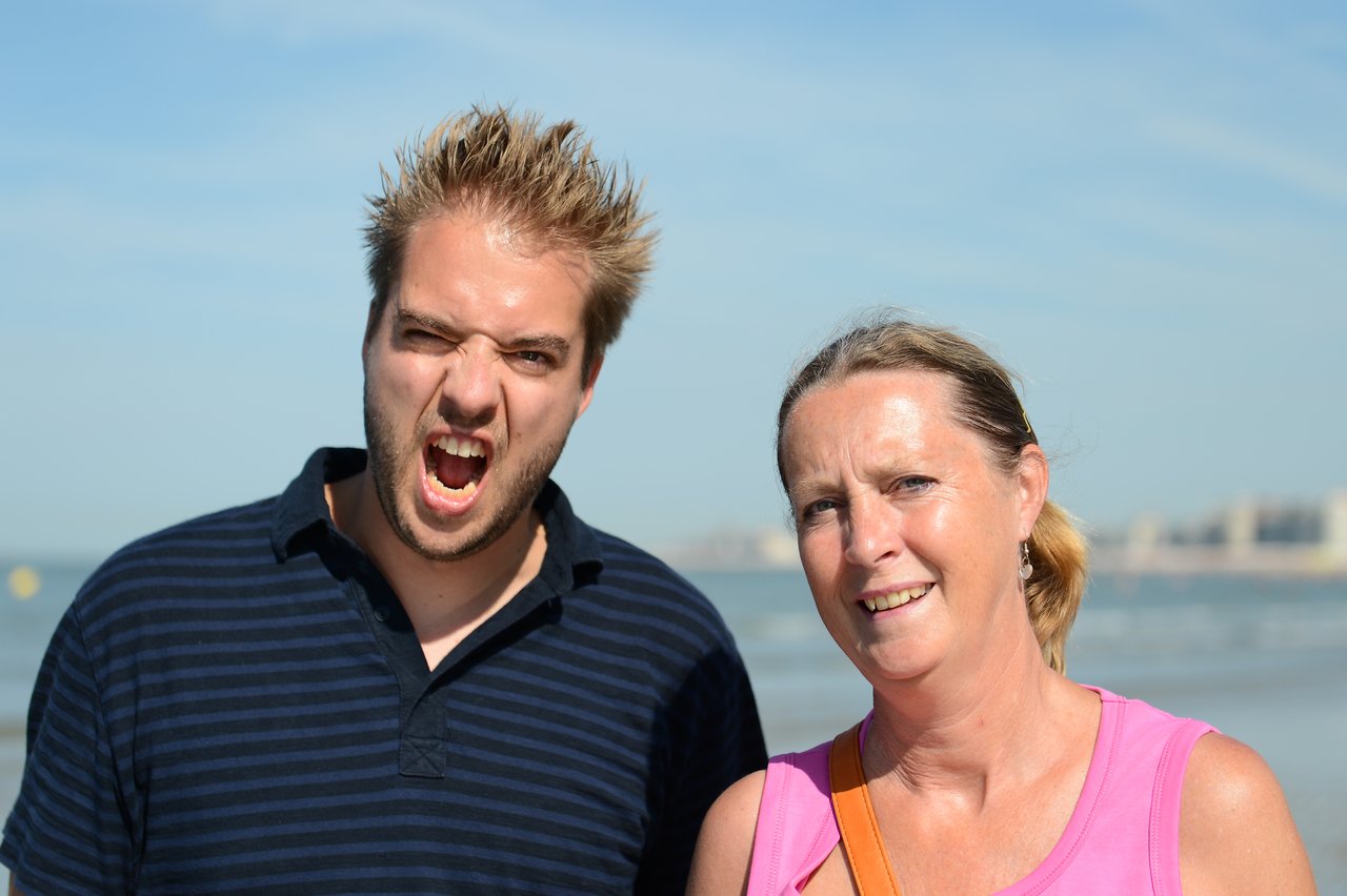 A young man makes a playful face while standing next to a smiling woman on a sunny beach.