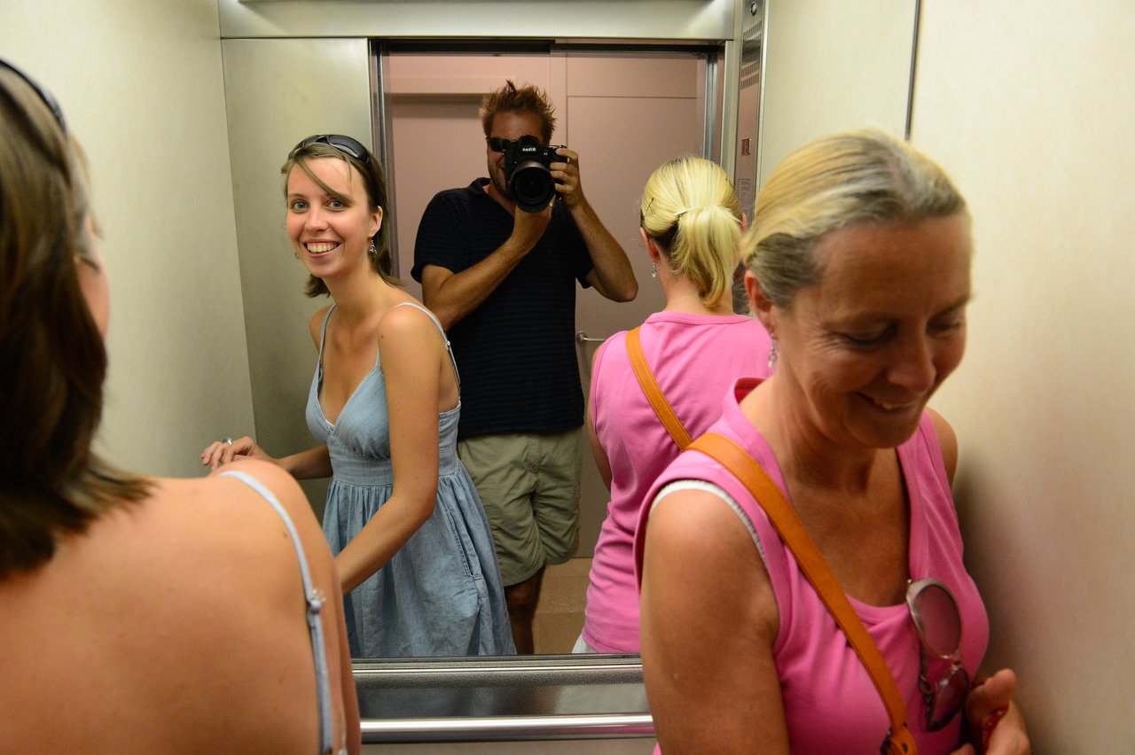 A man takes a photo in an elevator mirror while three women stand and smile in different directions.