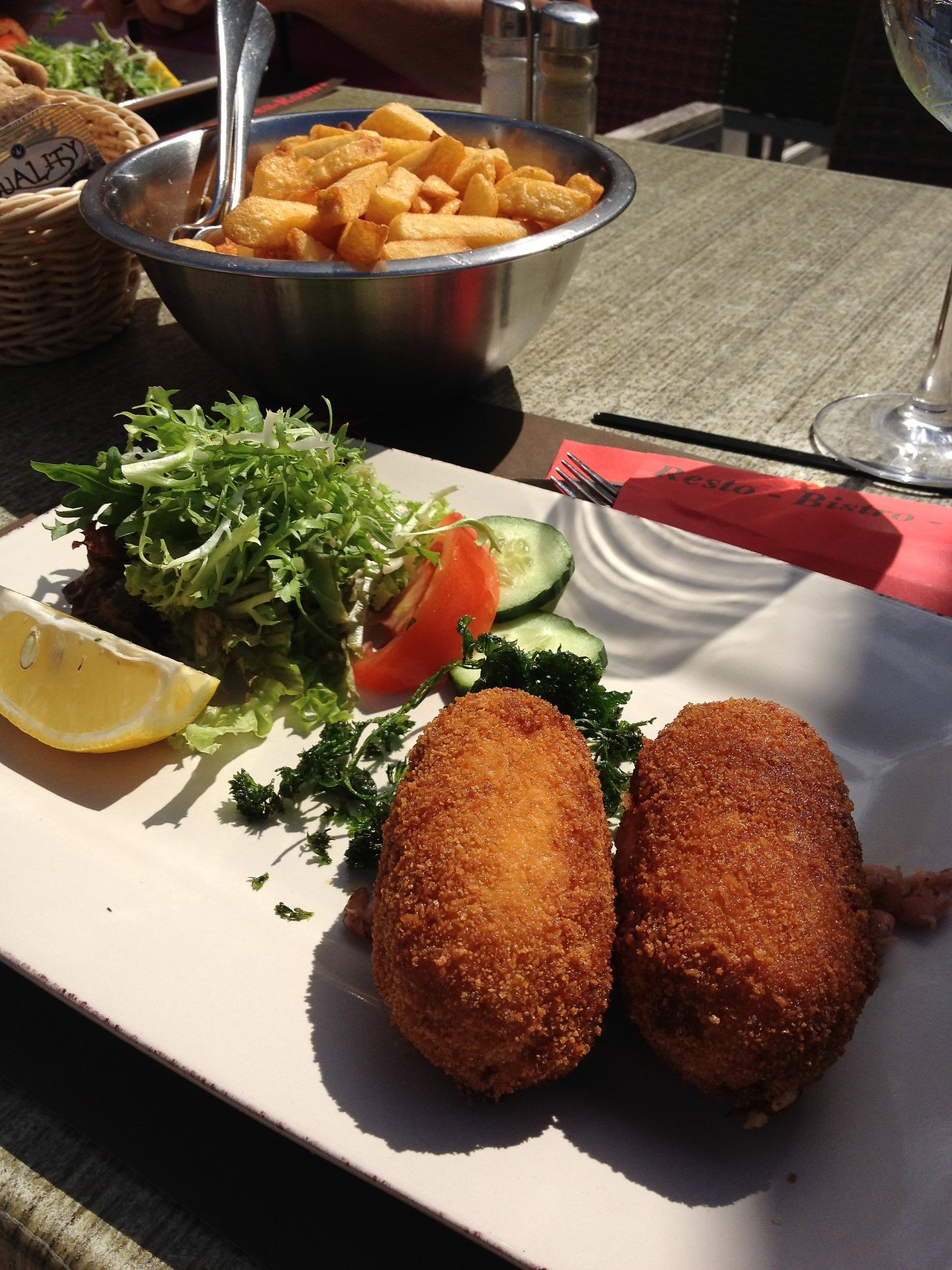 Two crispy shrimp croquettes on a plate with salad, lemon, and parsley, served alongside a bowl of fries.