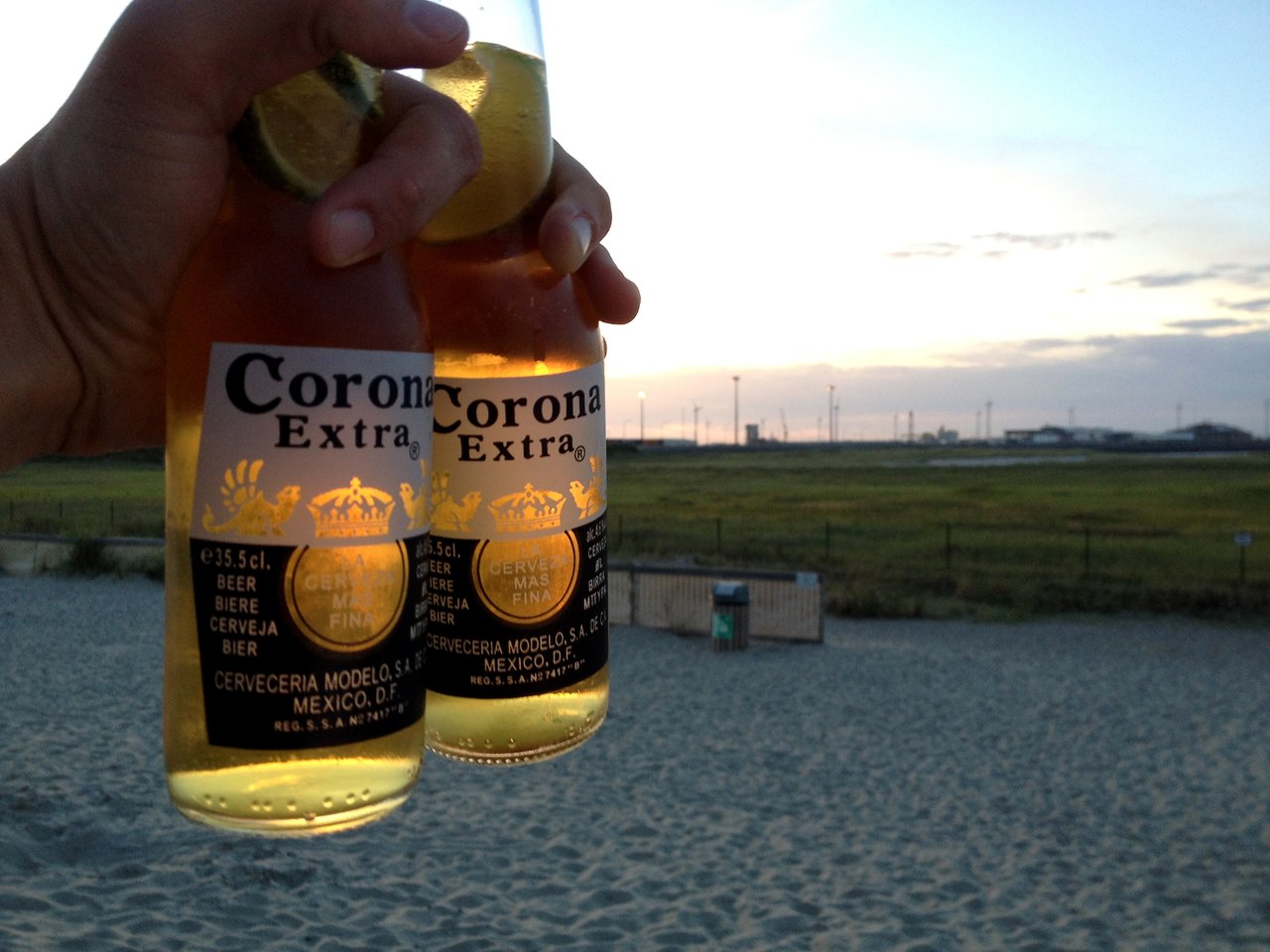 A hand holds two bottles of Corona beer with lime wedges, overlooking a sandy beach at sunset.