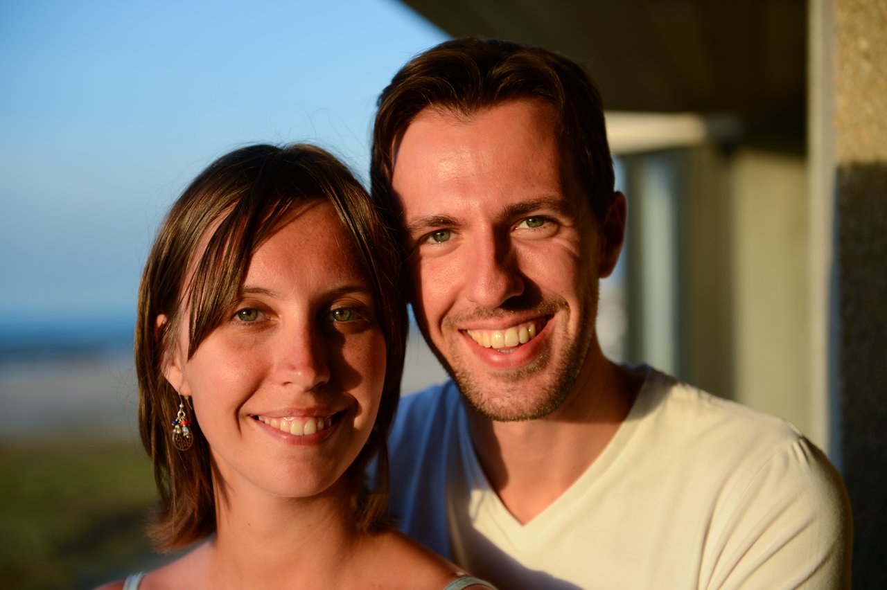 A smiling couple poses closely together in warm sunlight, looking at the camera.