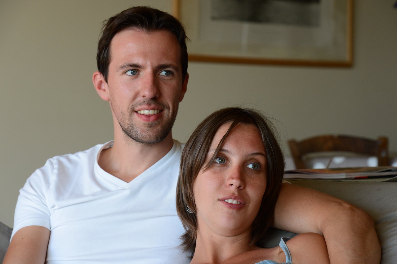 A man and woman sit closely on a couch, looking in the same direction with relaxed expressions.