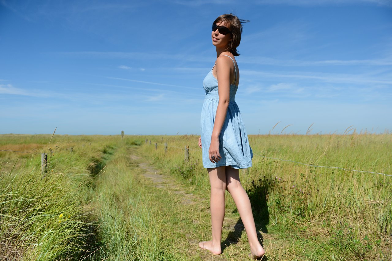 A woman in a light blue dress and sunglasses walks barefoot on a grassy path under a clear sky.