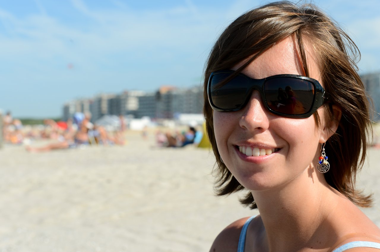 A woman wearing sunglasses and earrings smiles at the camera while sitting on a sandy beach.