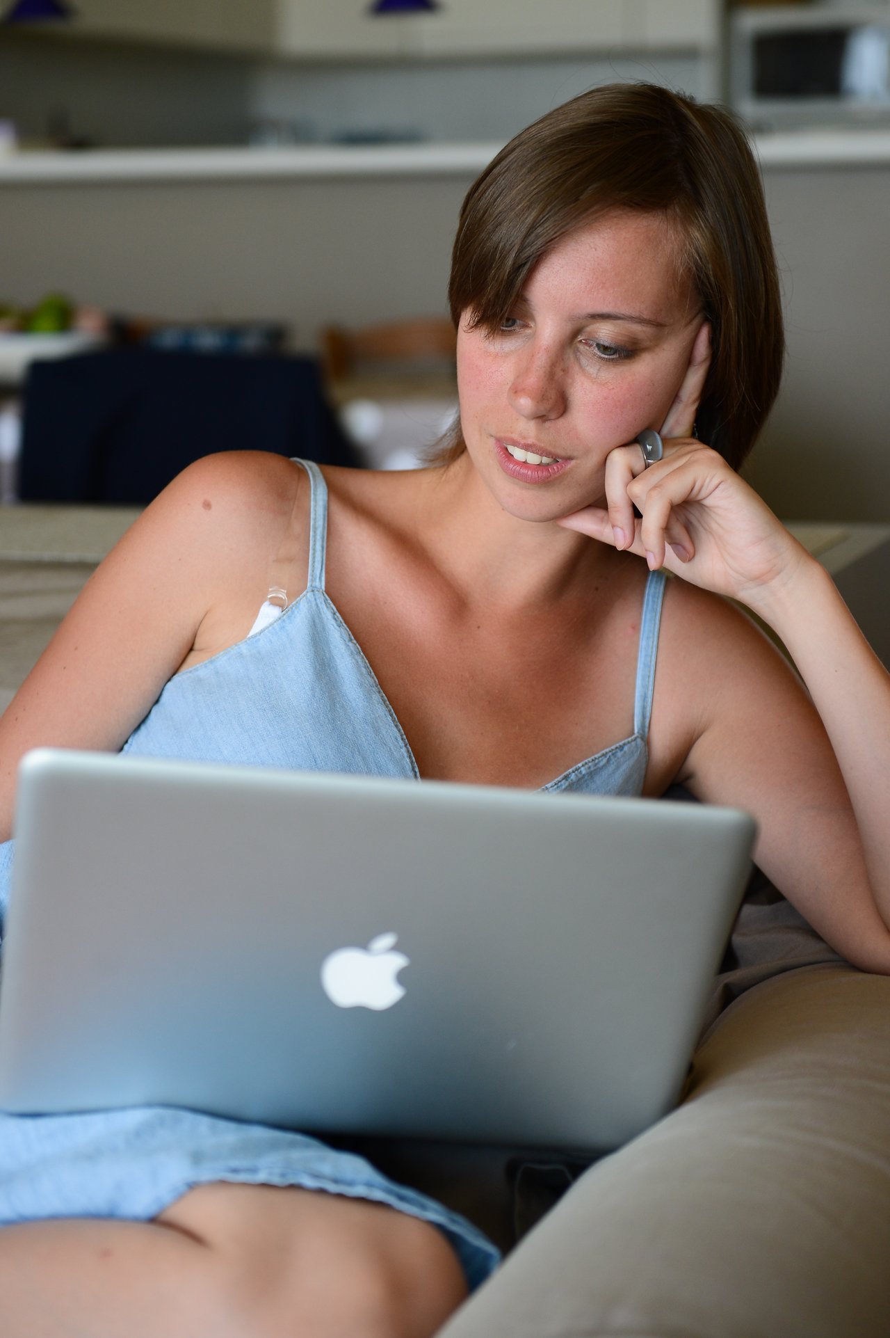 A woman sits on a couch, focused on her laptop screen while resting her head on her hand.